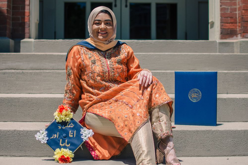 Graduate in orange traditional dress sits on steps with diploma and decorative bag.