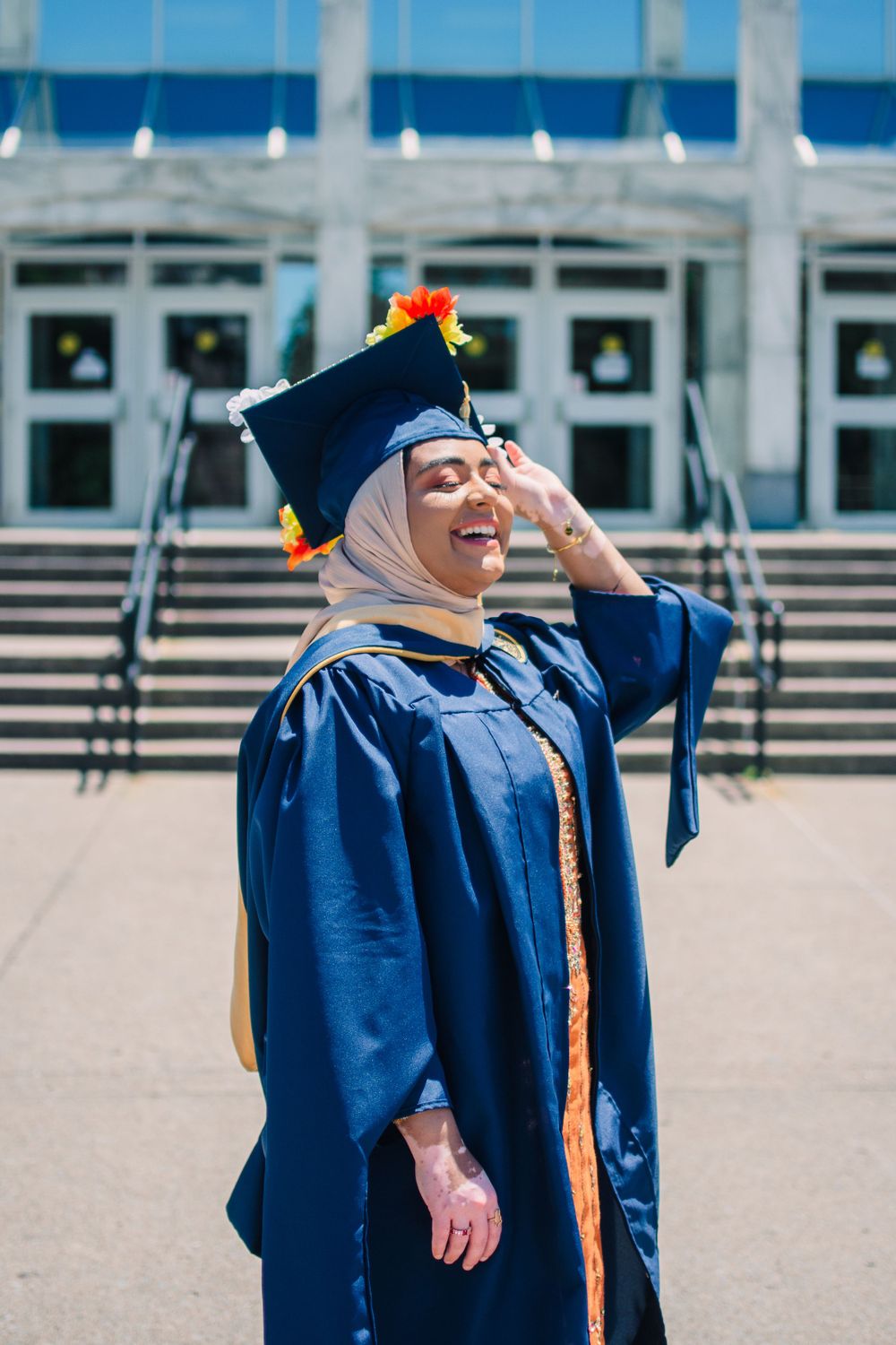 Graduate in blue cap and gown laughing joyfully on campus steps.