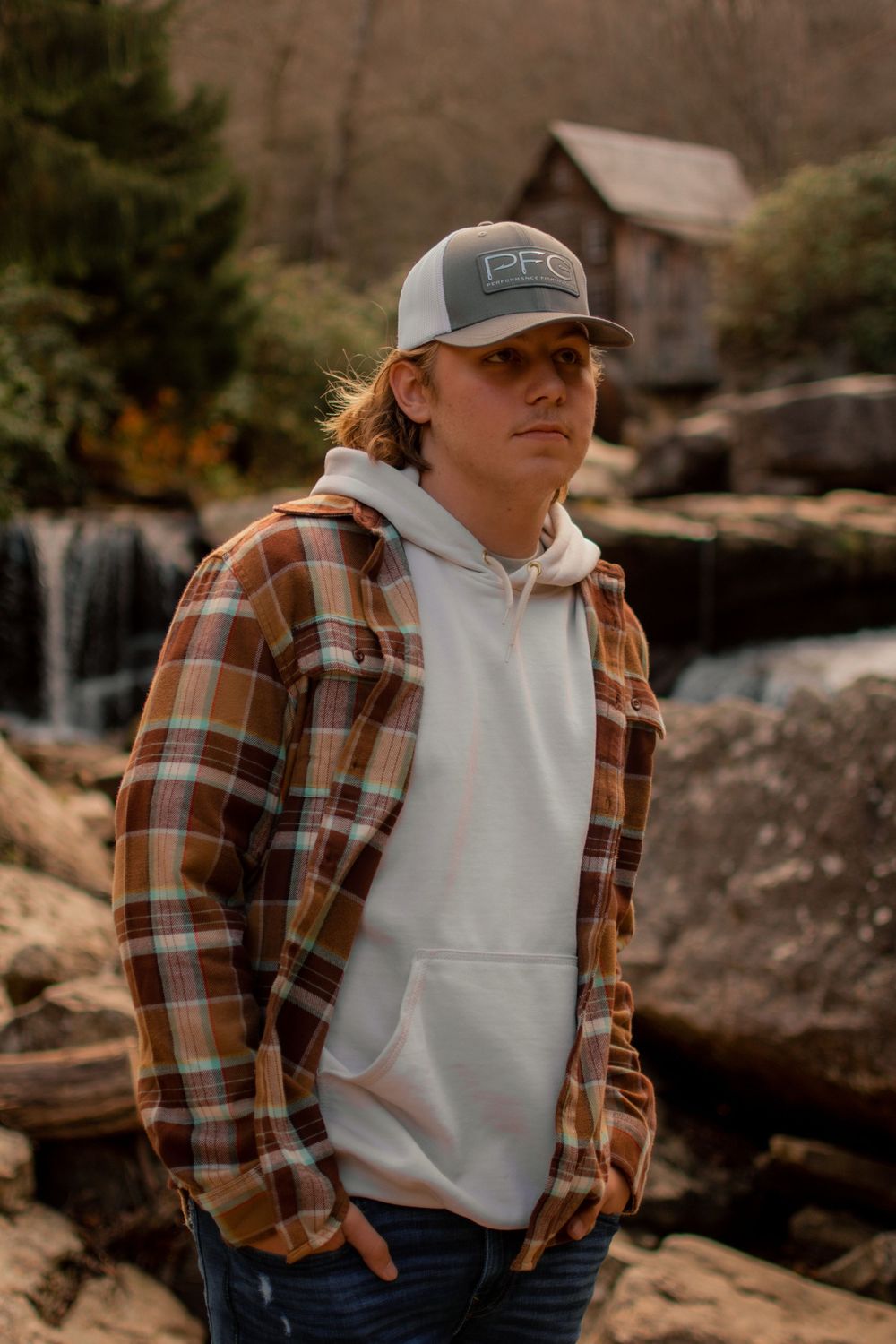 Person wearing plaid flannel shirt and white hoodie poses against rocky outdoor backdrop.