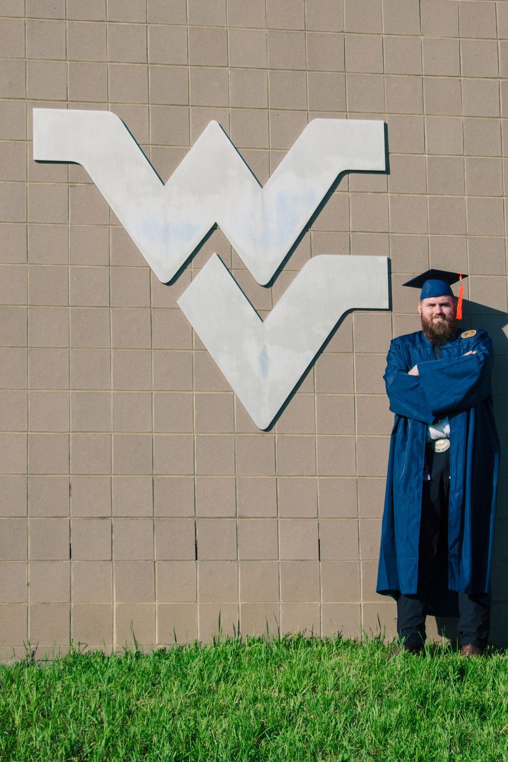 Graduate in cap and gown poses against wall with large university letter.