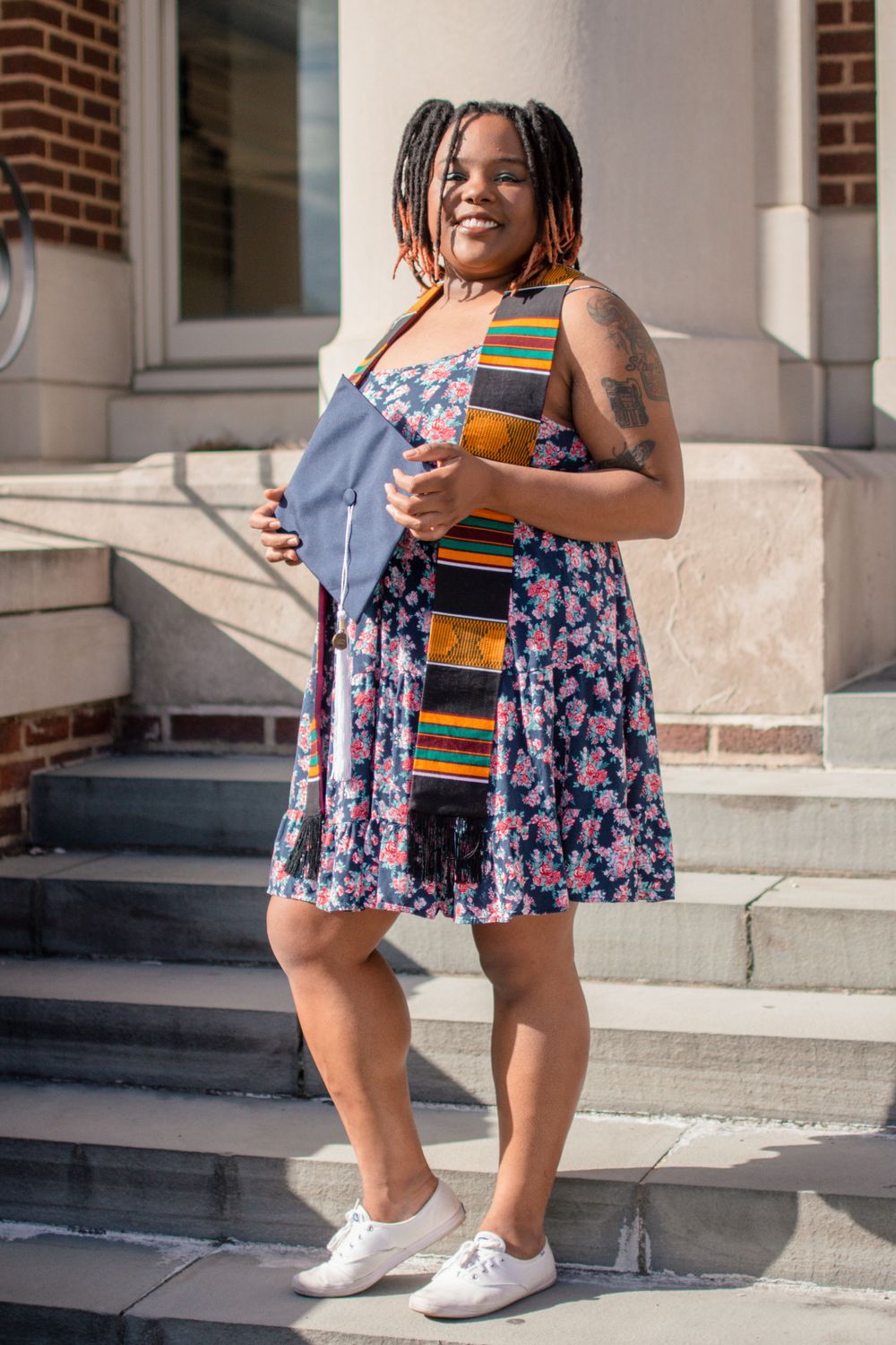 Graduate in floral dress and colorful stole stands on campus steps.