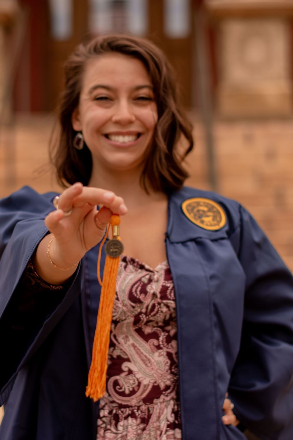 Graduate holding orange tassel in navy blue academic regalia with decorative patches and floral dress.