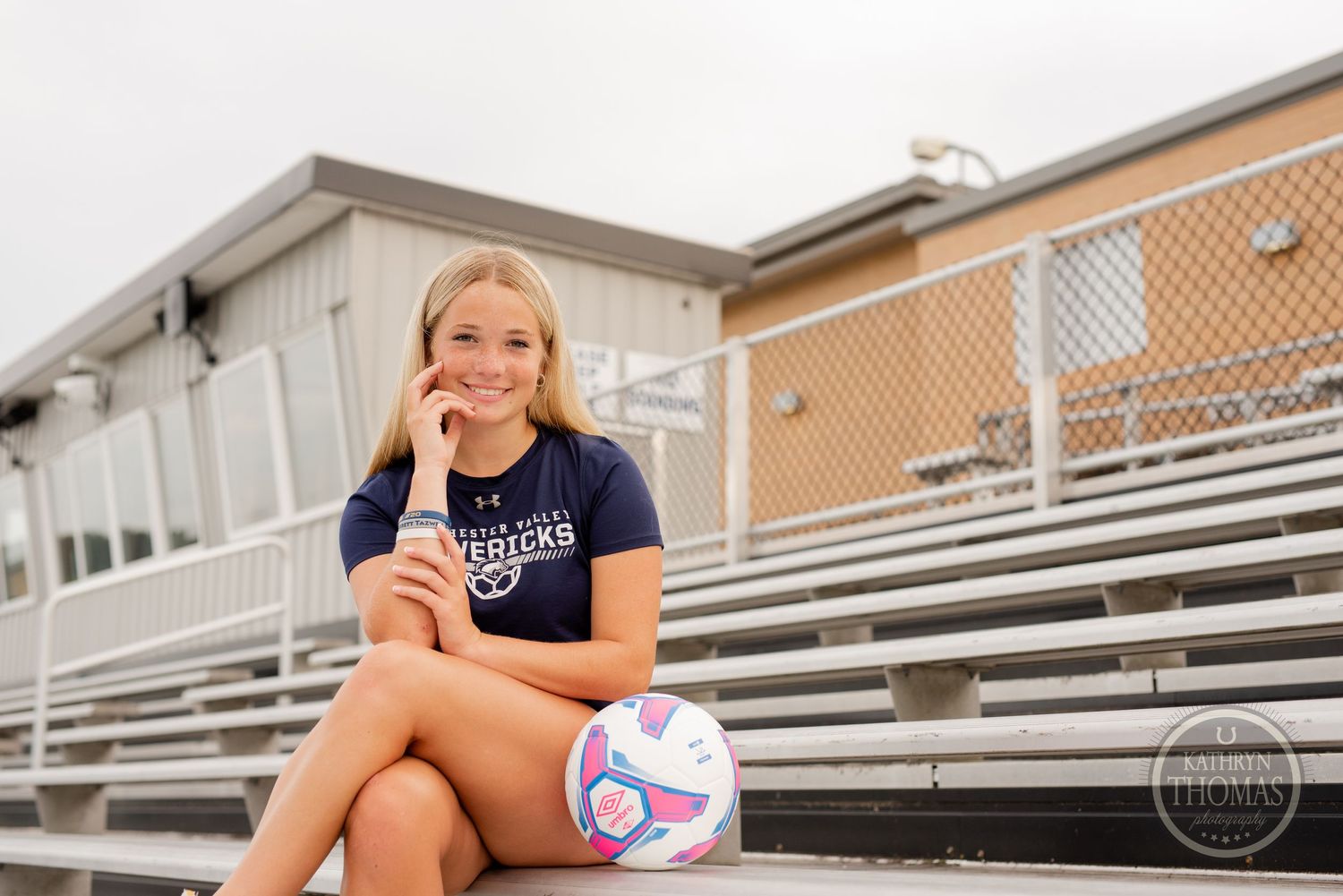 Soccer player sitting casually on metal bleachers with ball during outdoor photo session.