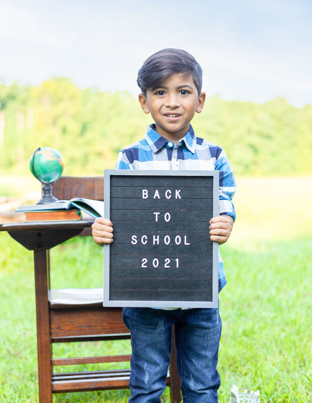 Student holds a letterboard sign reading 'Back to School 2021' with a decorative globe on a wooden desk outdoors.