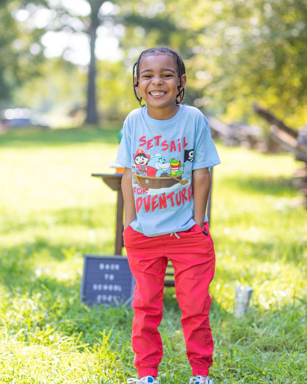 Student in blue shirt and red pants smiles while standing in a sunny outdoor setting.