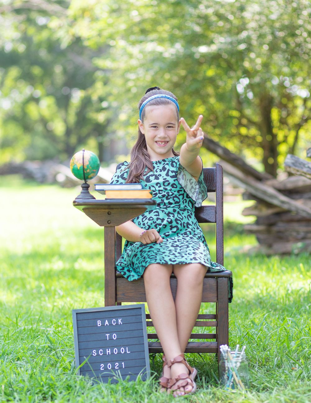 Student in floral dress poses with peace sign on wooden chair with globe and back-to-school sign.