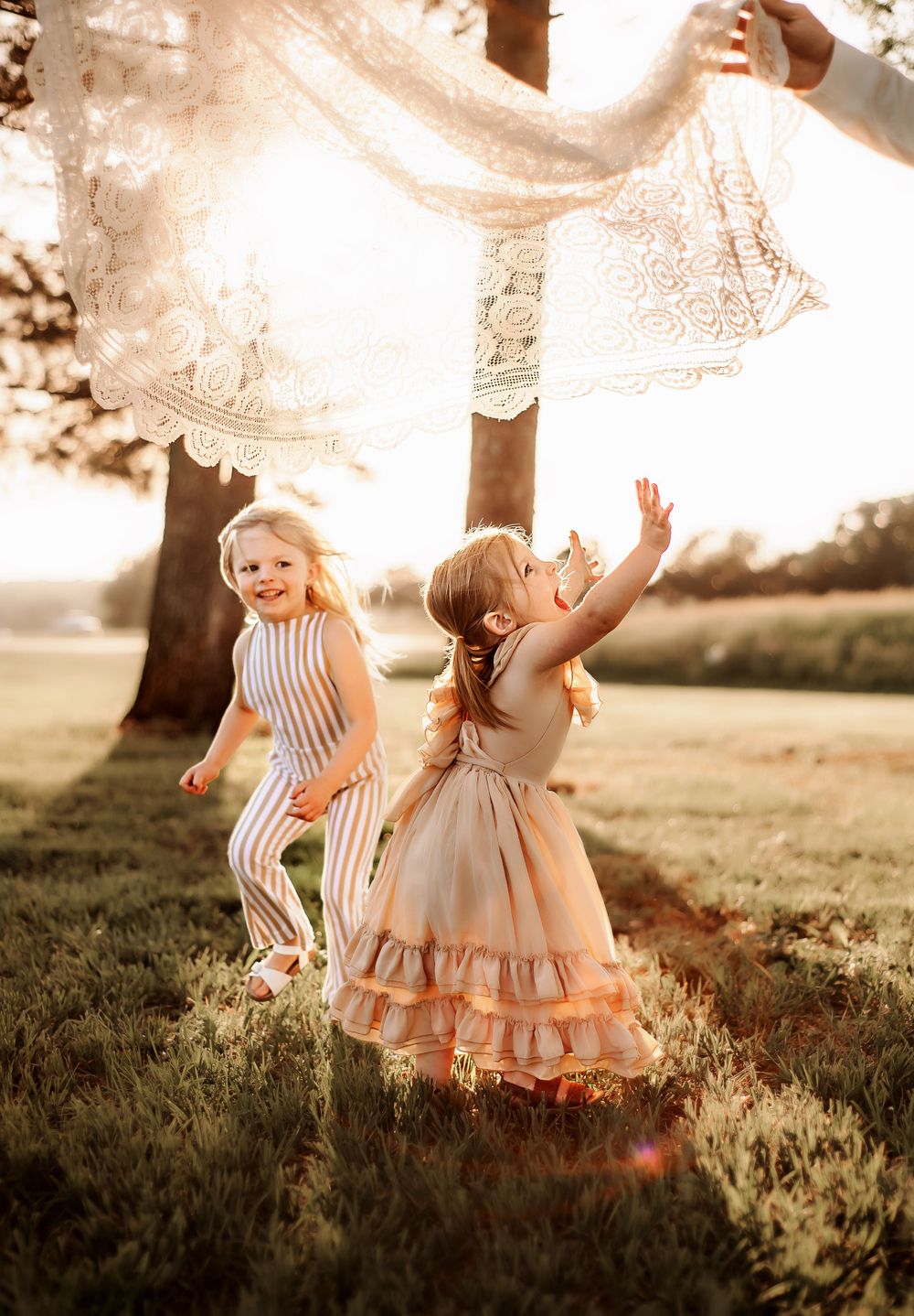 Two children playing under flowing fabric in a sunlit field wearing vintage-style clothing.