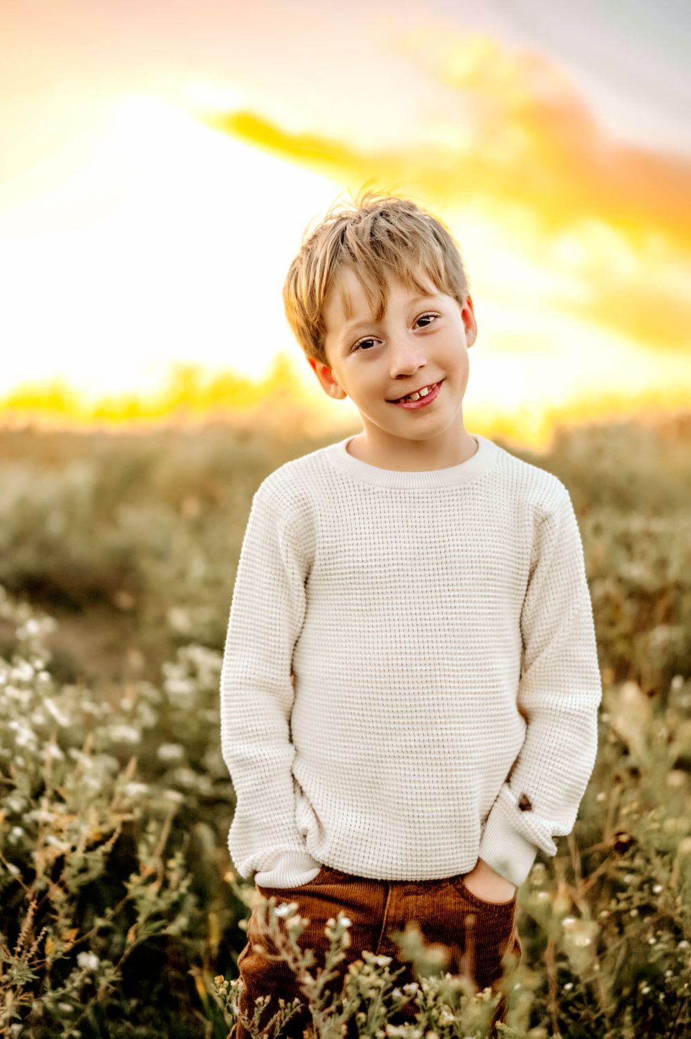 A young one smiles warmly while standing in a field at sunset wearing a cream knit sweater.