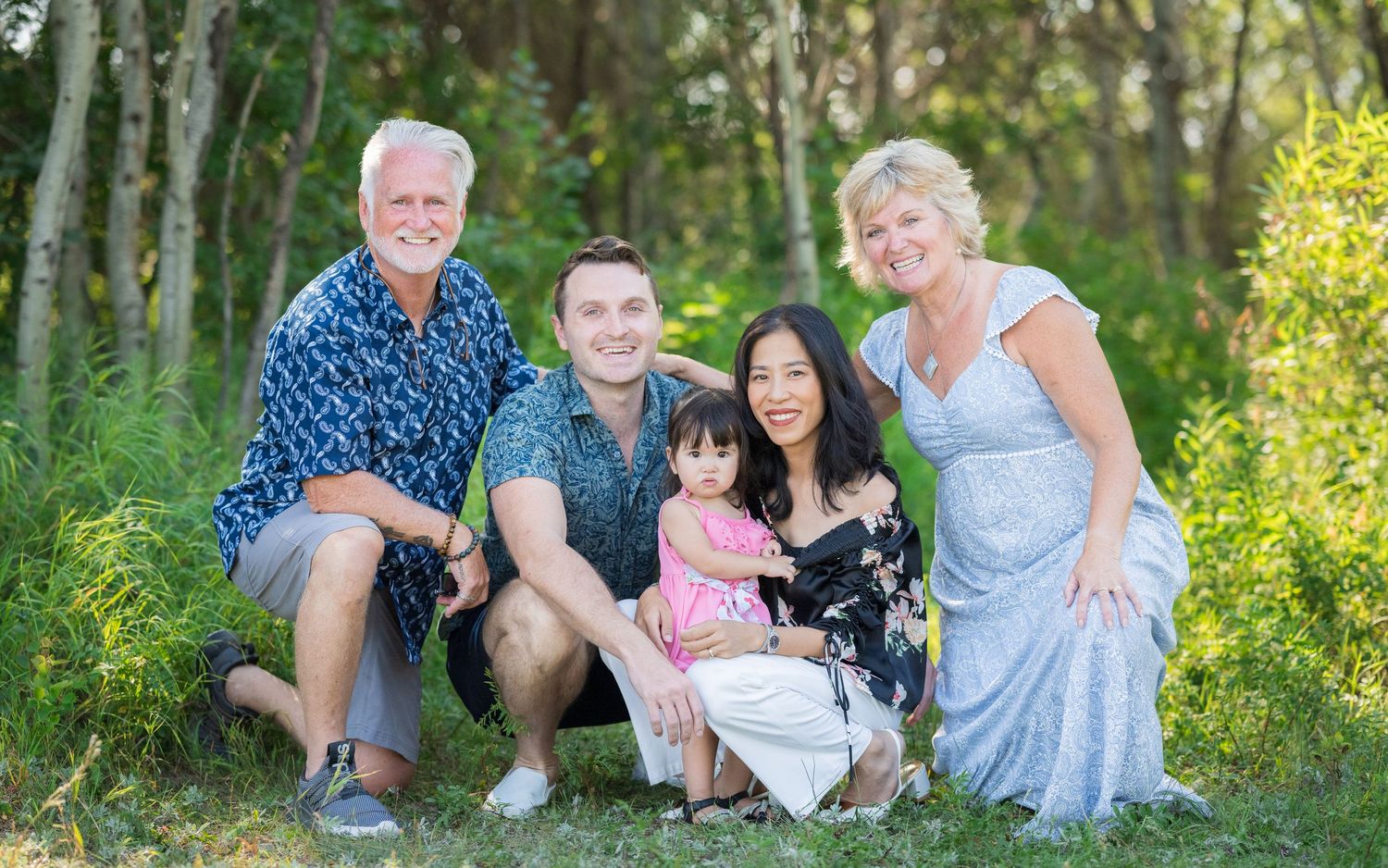Multi-generational family portrait taken outdoors in a sunny wooded setting.