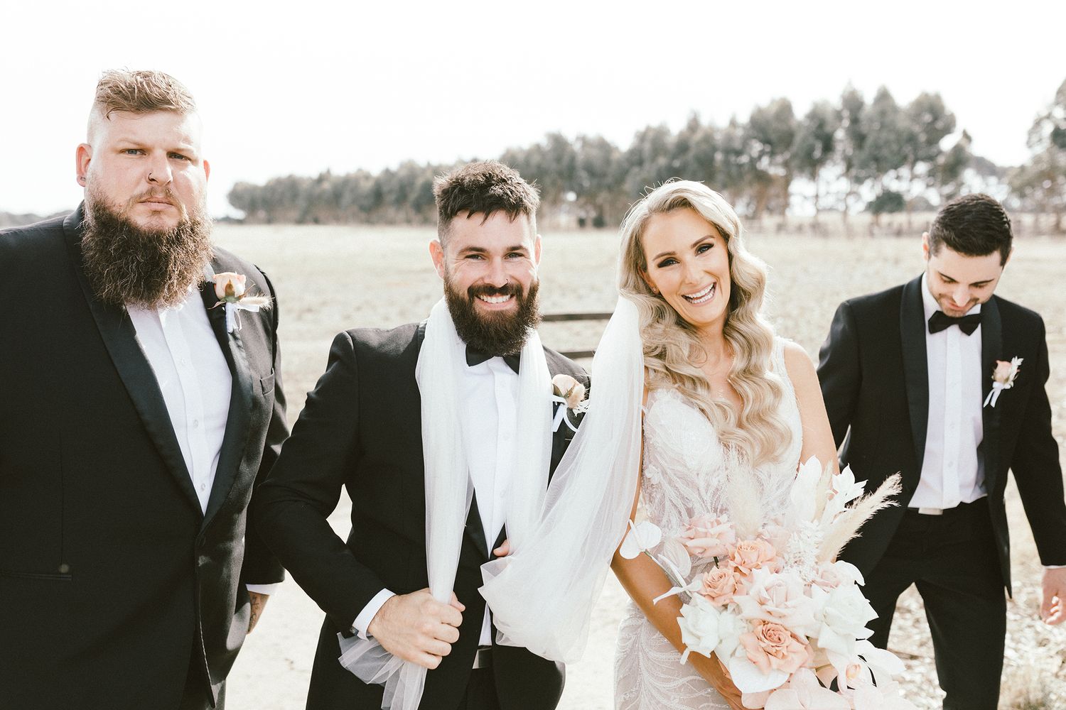 Groomsmen in black tuxedos standing together outdoors at a rustic wedding venue.