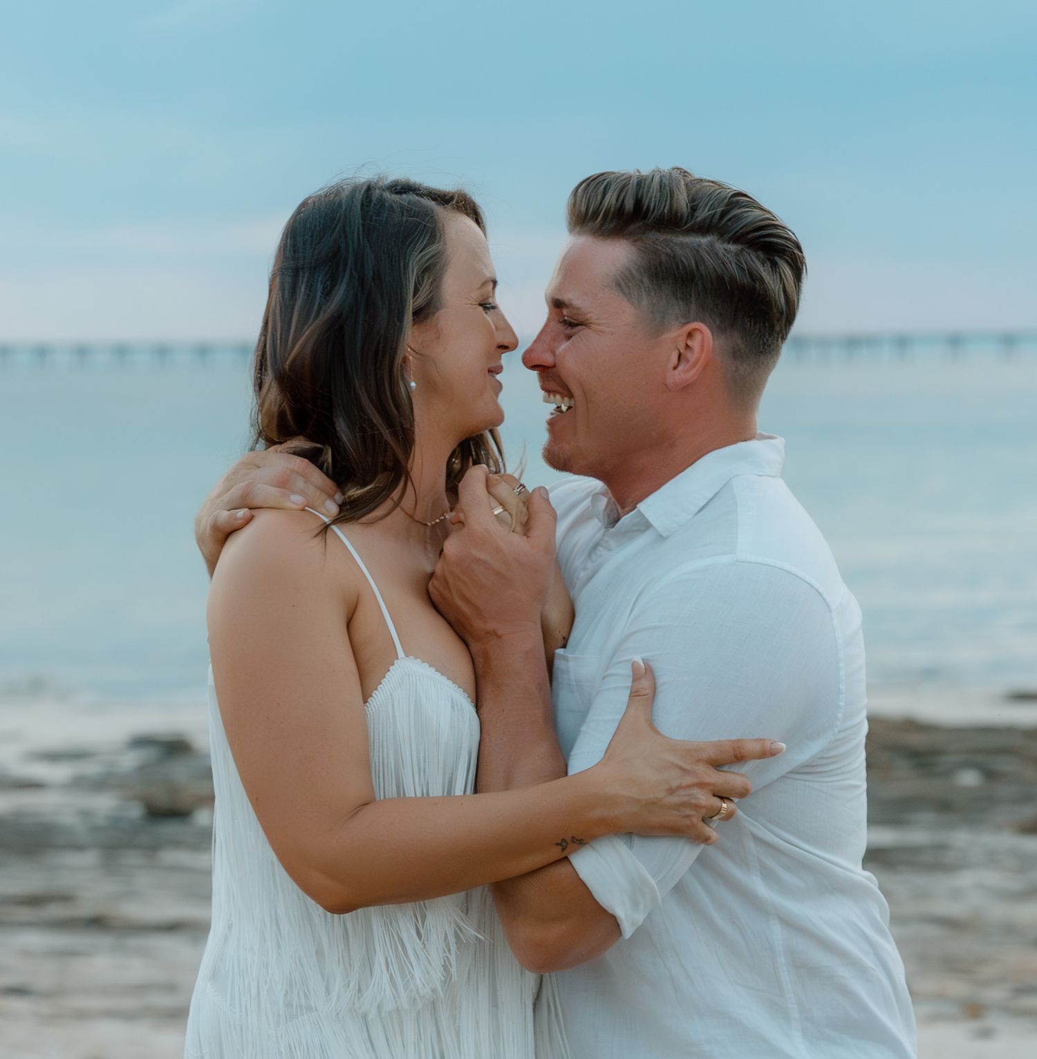 Romantic couple sharing an intimate moment on a beach with a pier in the background.