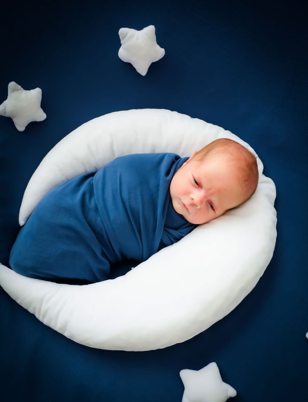 Sleeping infant wrapped in blue swaddle on curved white pillow against dark backdrop.