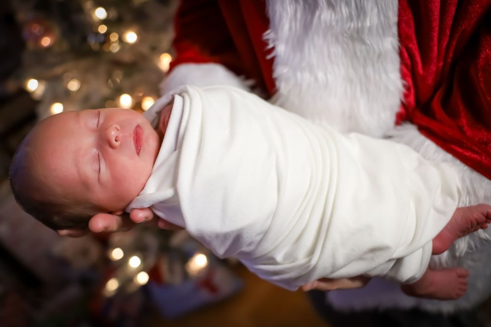 A baby wrapped in white against red fabric with twinkling lights in the background.