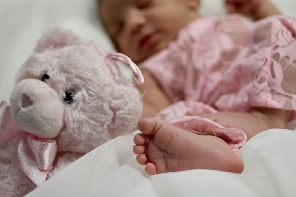 Tiny baby feet next to a soft pink plush toy on white bedding.