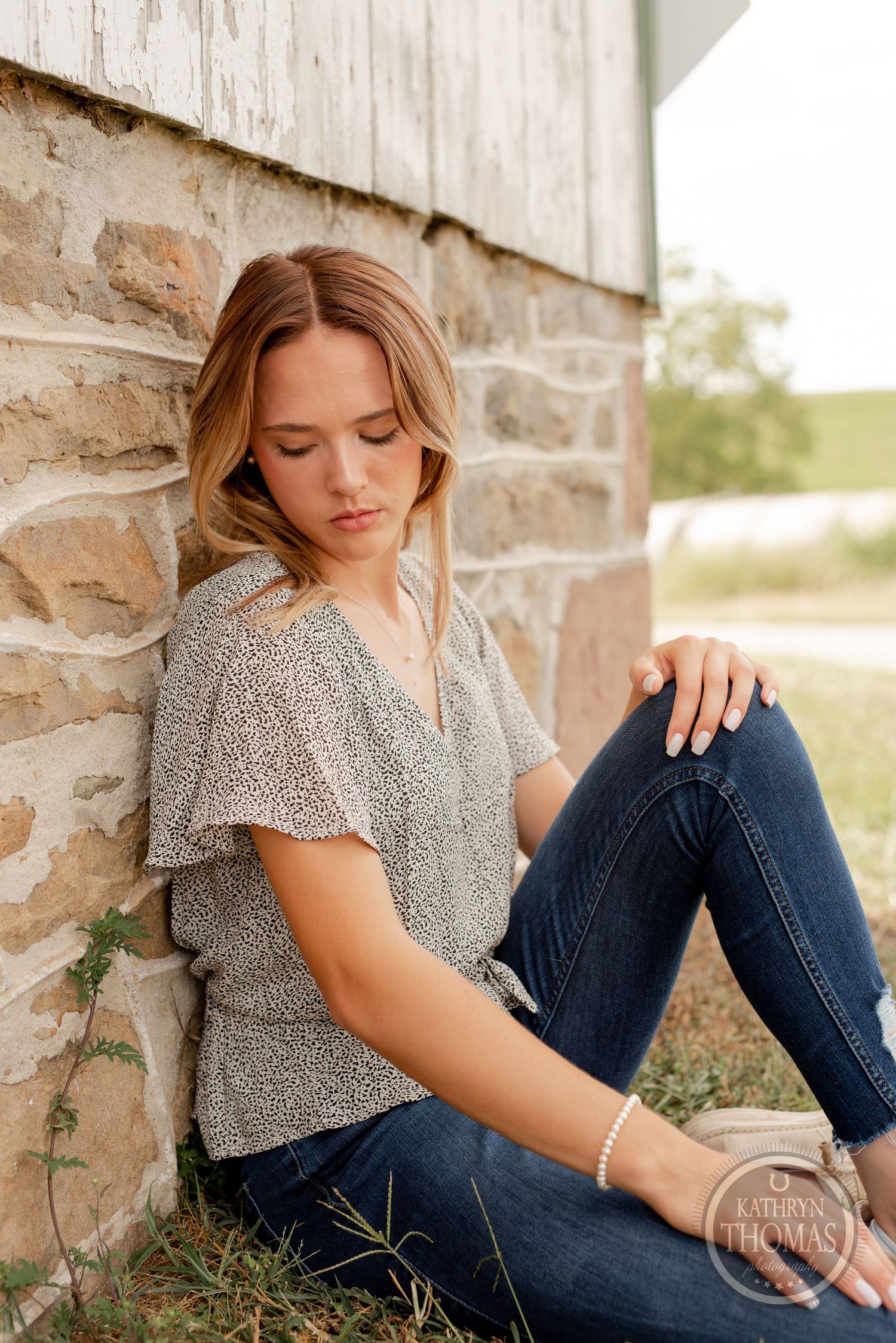 A person in a floral top and jeans sits casually against a stone wall in a countryside setting.