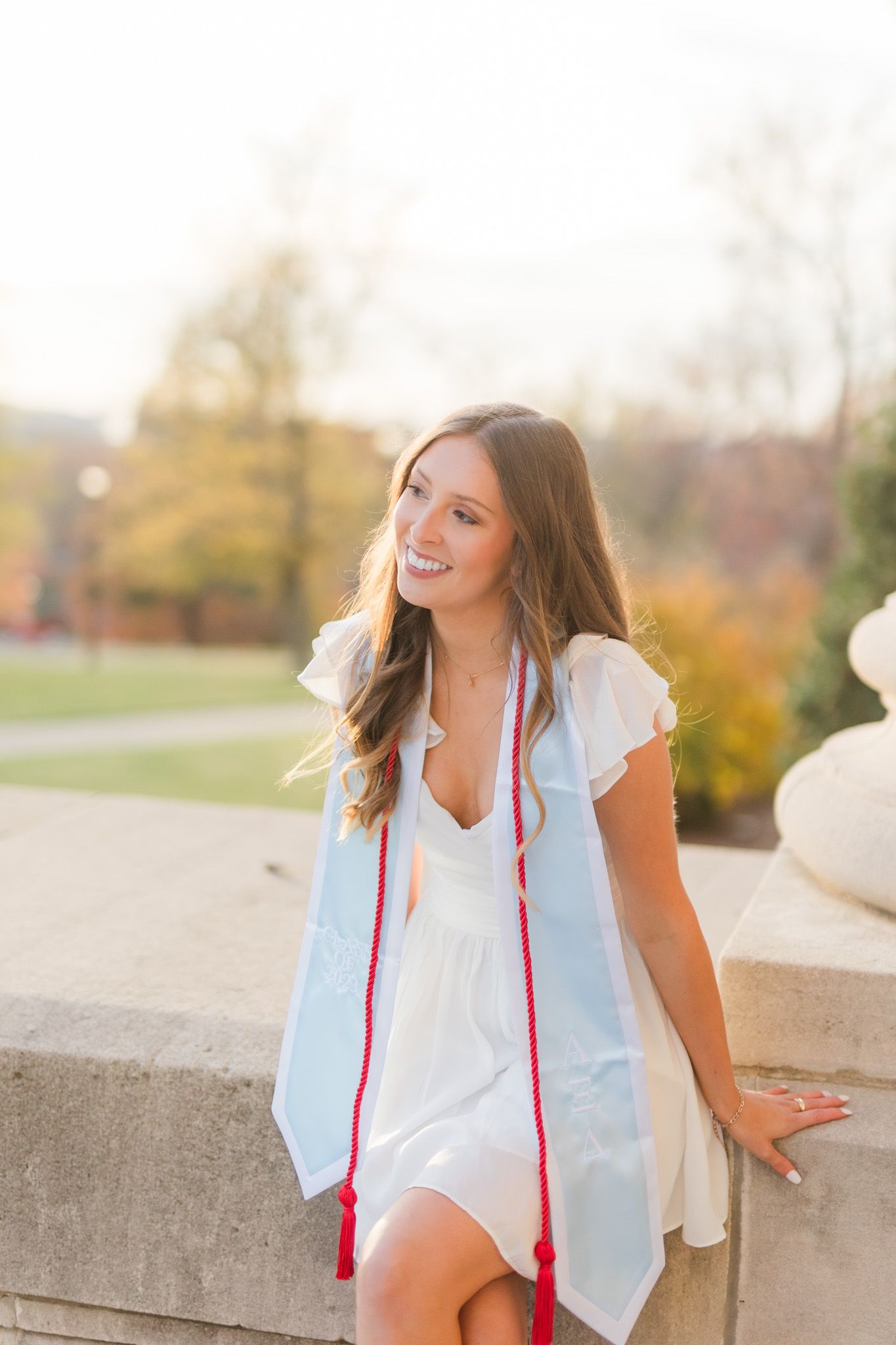 Graduate in white dress poses against stone wall during golden hour photography session.