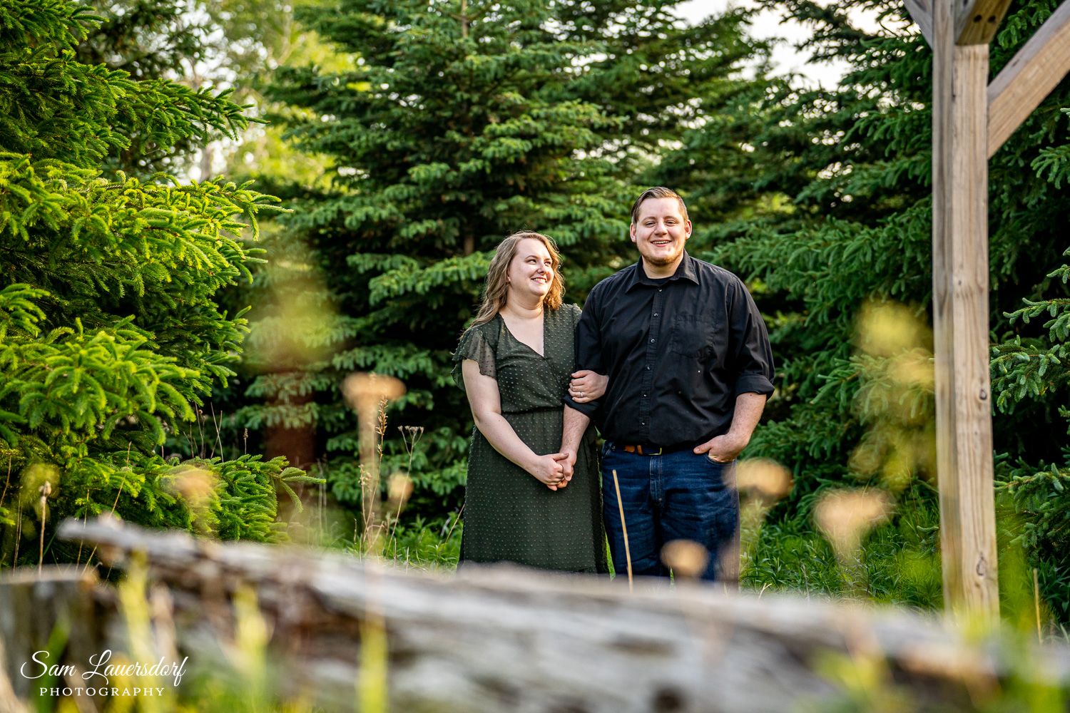 A cheerful couple poses together on a rustic wooden deck with evergreen trees in the background. Couples photography
