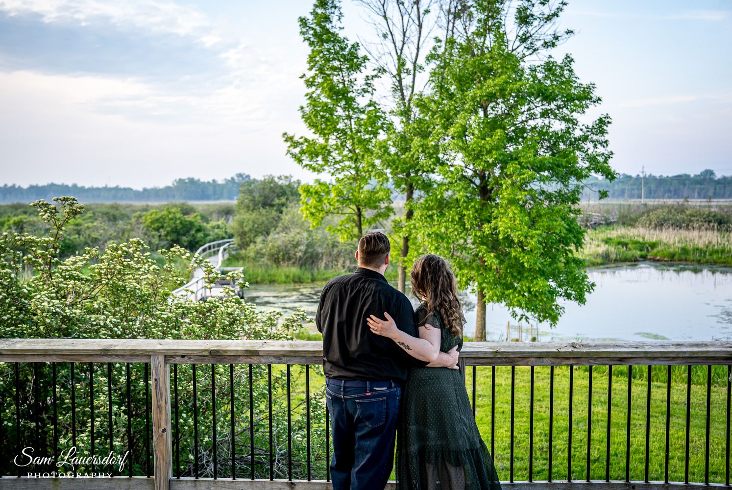 A couple stands together on a deck overlooking a serene lake and tree-lined landscape. creative couples photography