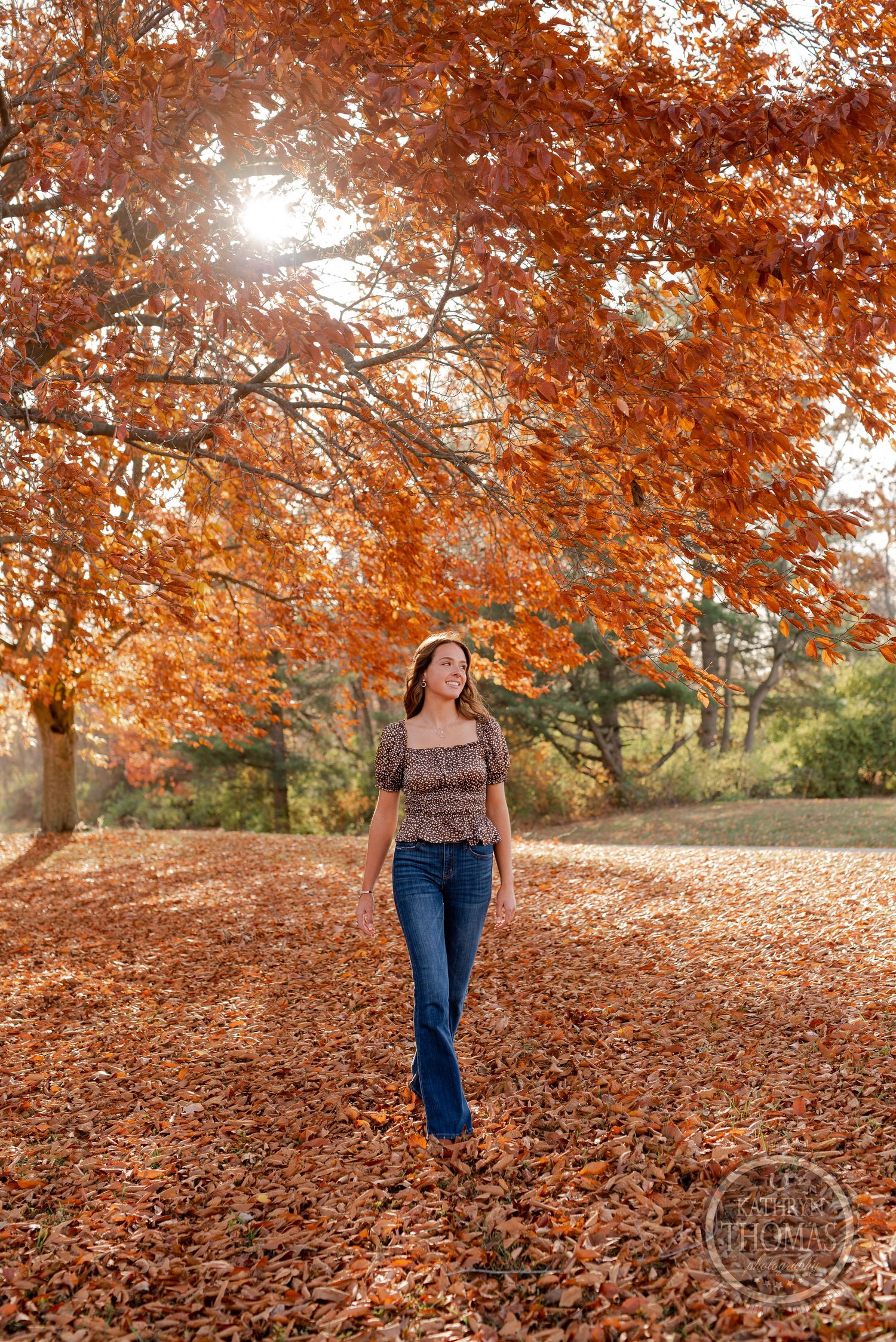 A casual fashion portrait of someone walking through fallen autumn leaves under orange trees.