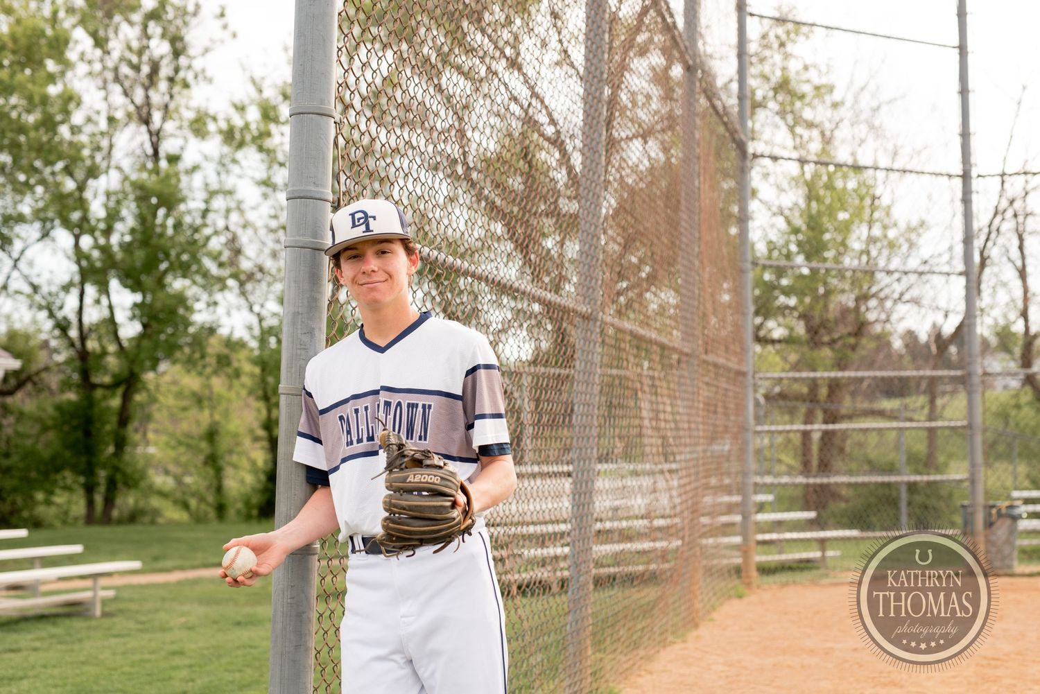 Baseball player in white uniform stands near chain-link fence on baseball field.