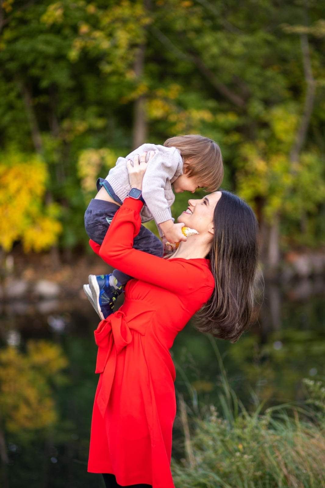 Woman in red dress lifts child playfully against autumn foliage background near water.