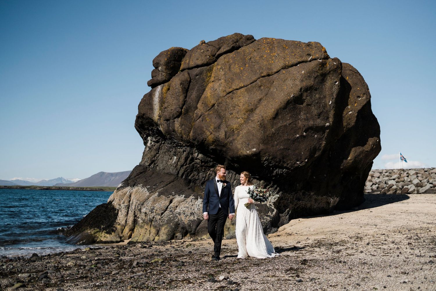 A couple in wedding attire poses in front of a massive boulder on a rocky beach with mountains in the background.