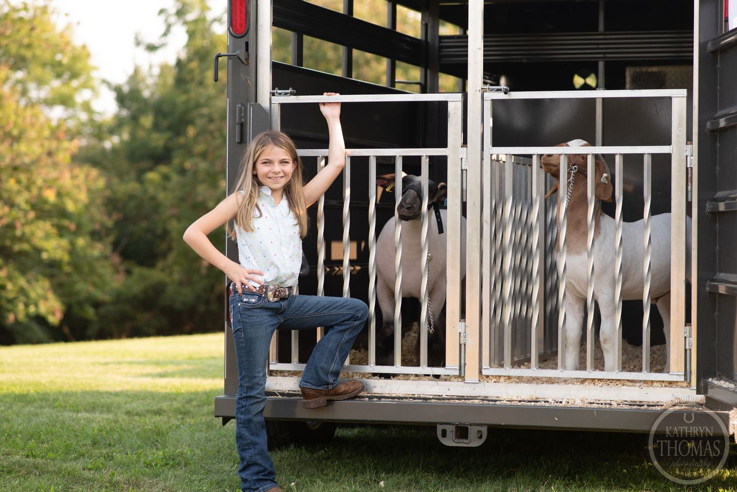 A livestock trailer stands in a field as someone poses confidently beside it.