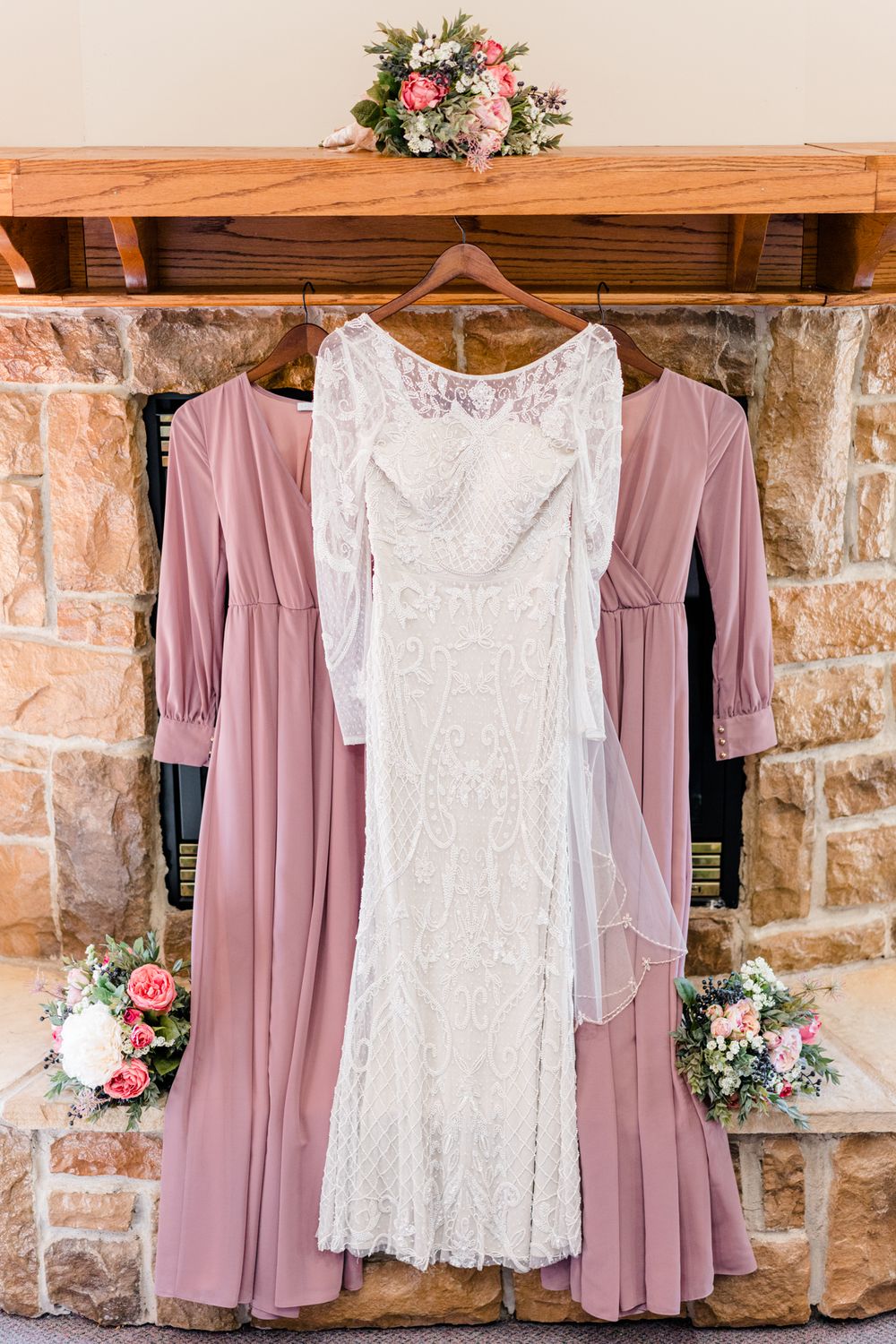 A white lace wedding dress and mauve bridesmaid dresses hanging against a stone wall with floral decorations.