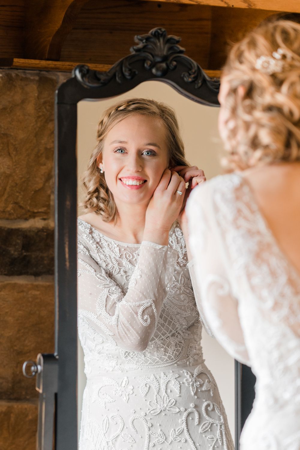 A bride in a lace wedding gown puts on earrings while looking in an ornate black-framed mirror.