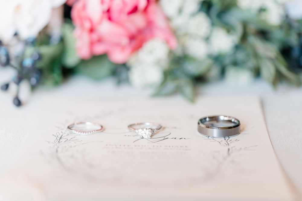 Three wedding rings arranged on white paper with pink flowers in soft focus.
