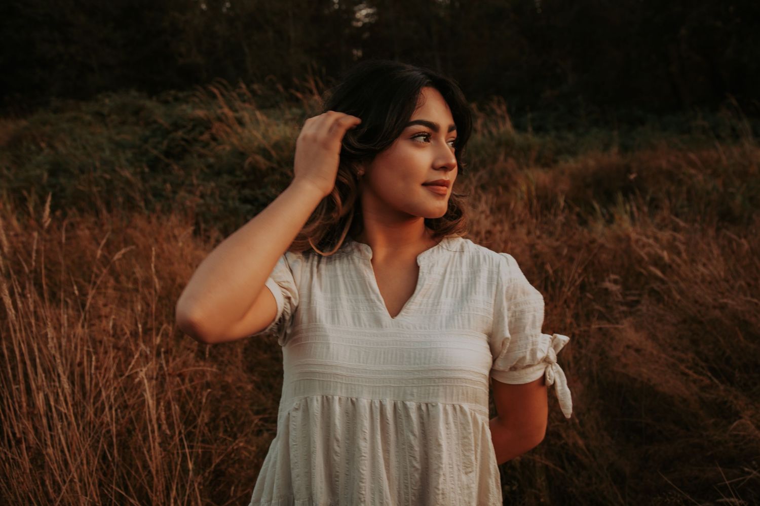 Young woman in a white dress touching her hair and gazing aside in a golden grassy field at sunset.