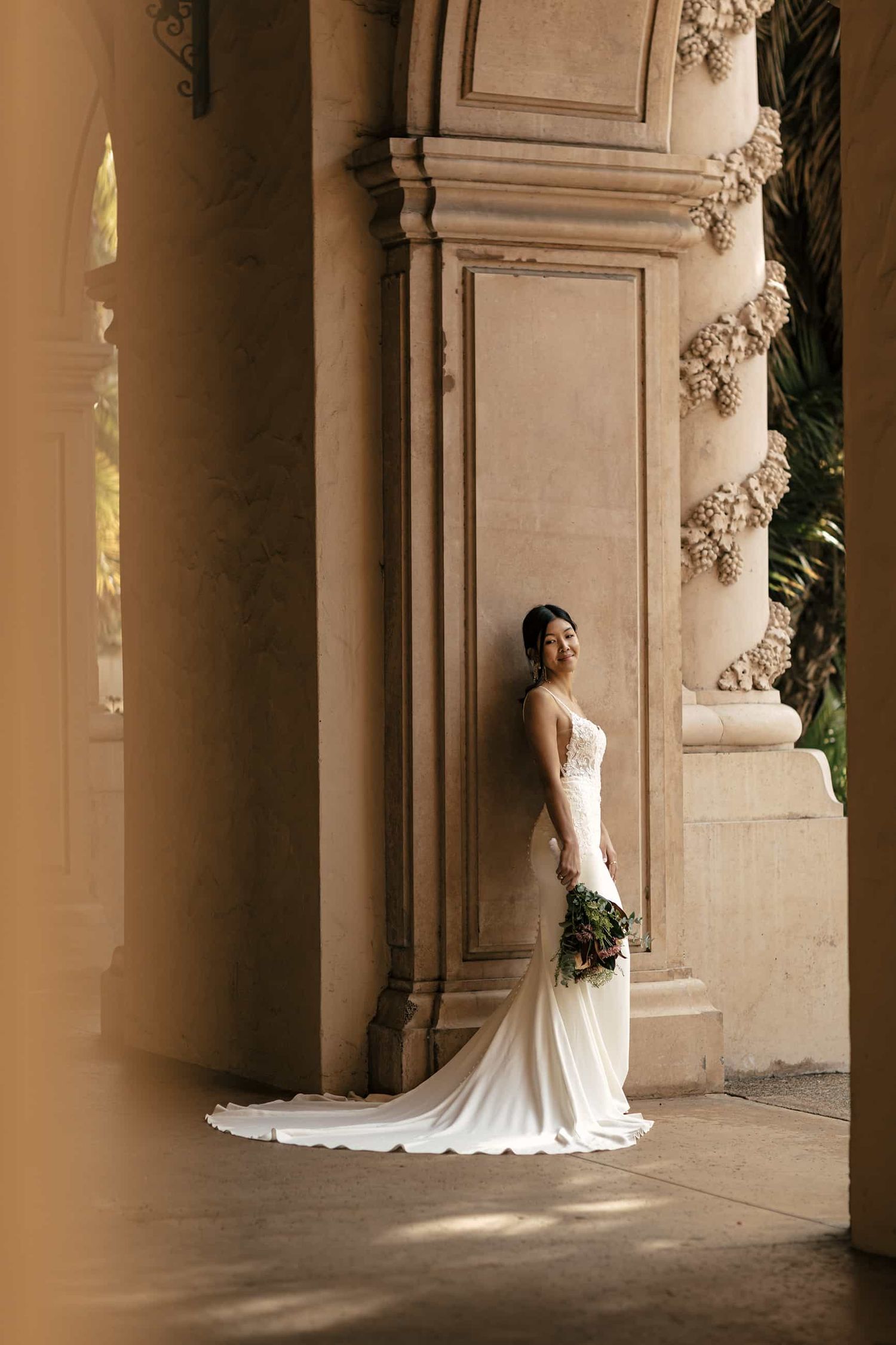 Elegant bride in fitted white gown poses beside ornate stone columns holding a bouquet.