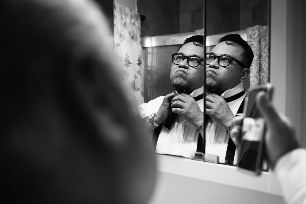 A frustrated groom frowns while tying his bow tie as father of the groom helps by playing a Youtube bow tie tutorial.
