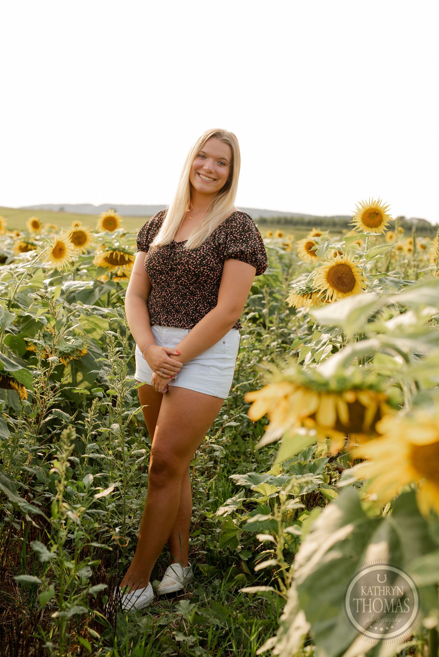 A person wearing a dark patterned top stands among tall blooming sunflowers during sunset.