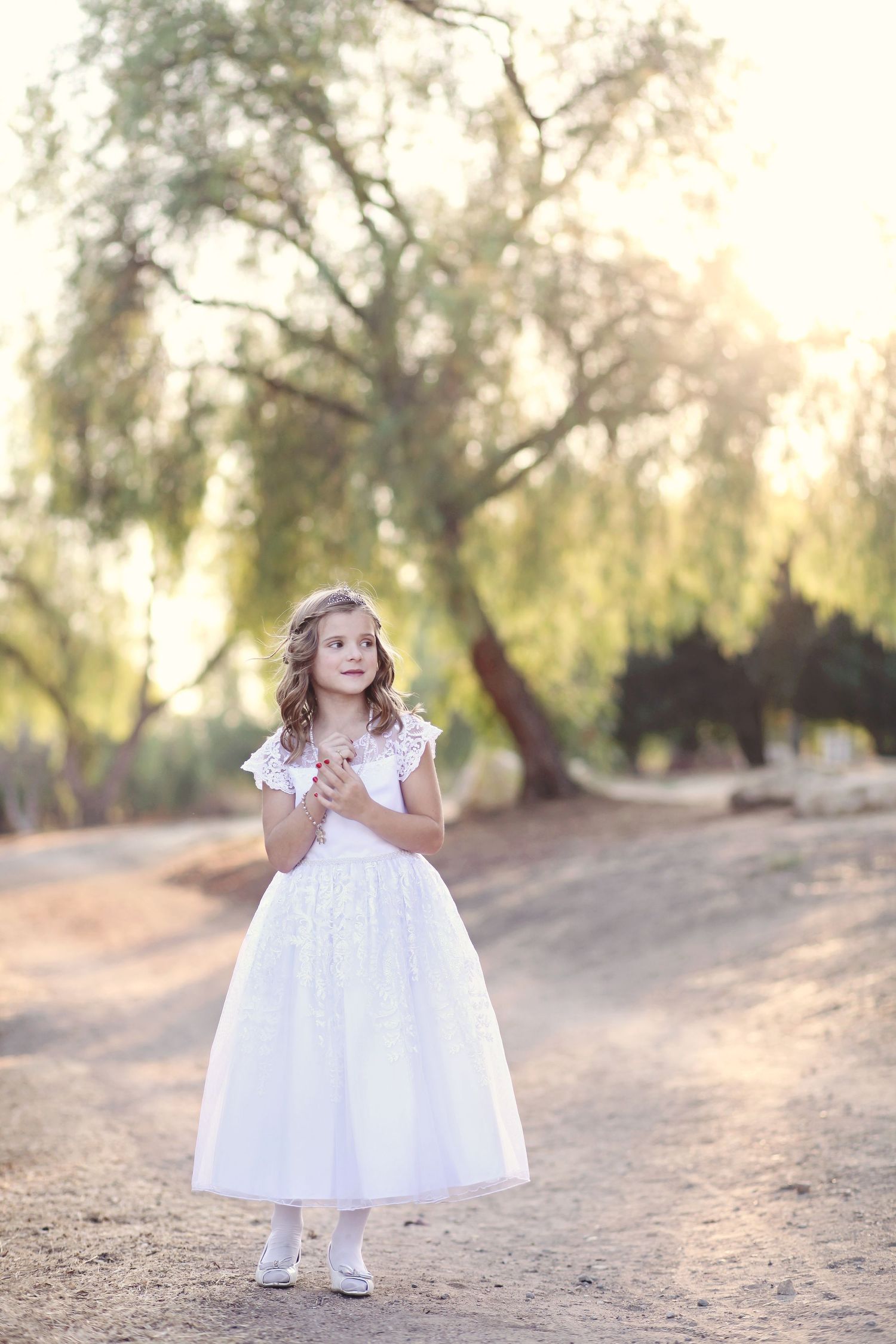 Young girl in white dress standing on dirt path under willow trees at sunset.