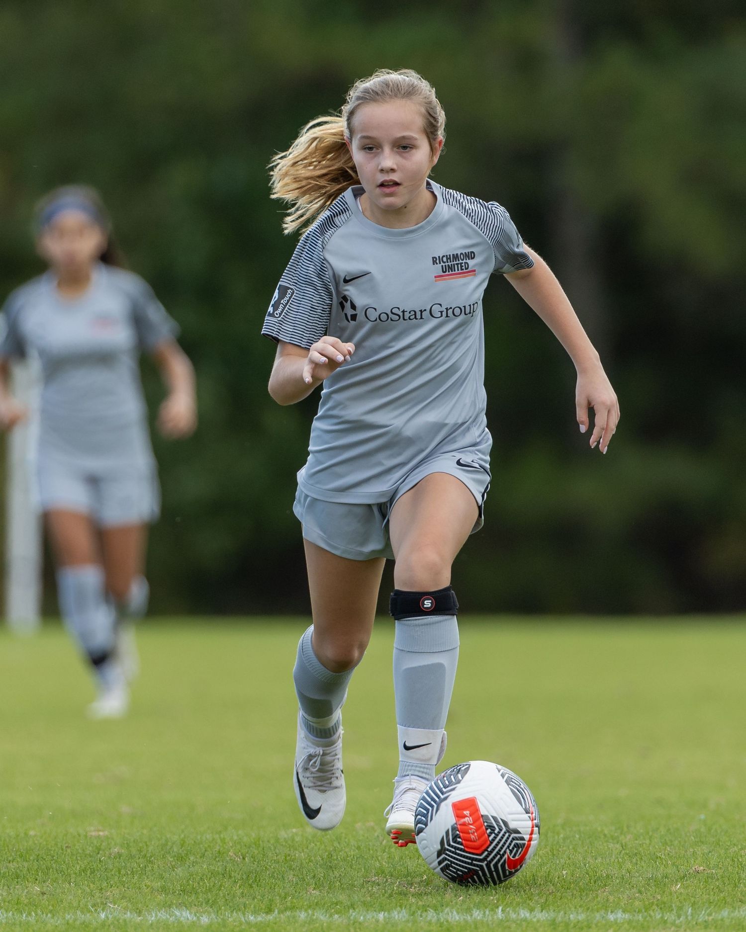 Soccer player in grey uniform dribbles ball across grass field during competitive match.