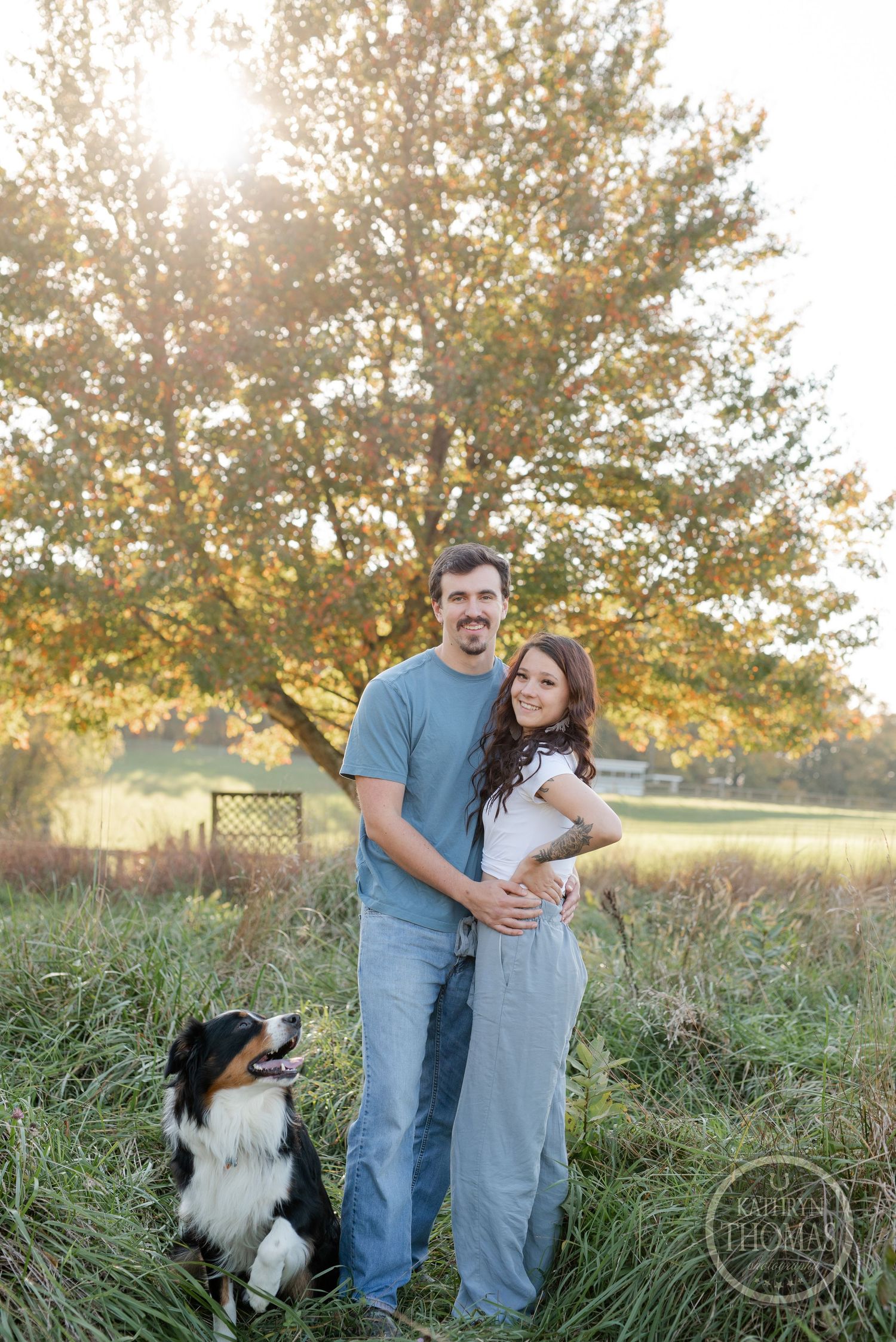 A couple stands together in a field at sunset with their attentive Border Collie companion.