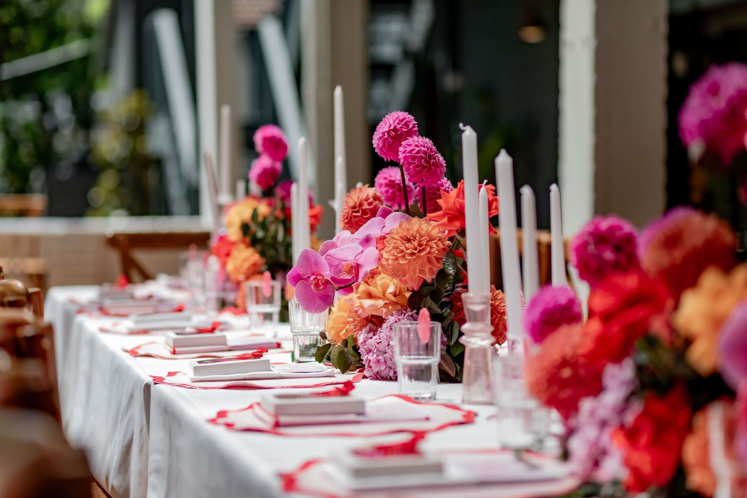 Long dining table decorated with pink and coral flowers, white candles, and red table settings for an elegant event.