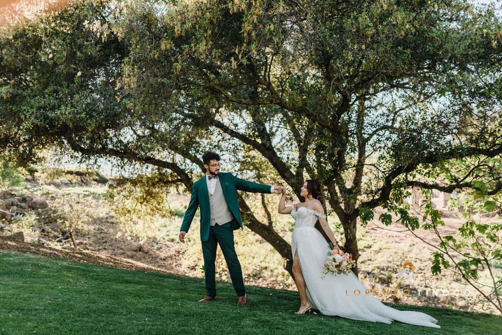 Wedding couple dances playfully on a green lawn beneath a large shady tree.