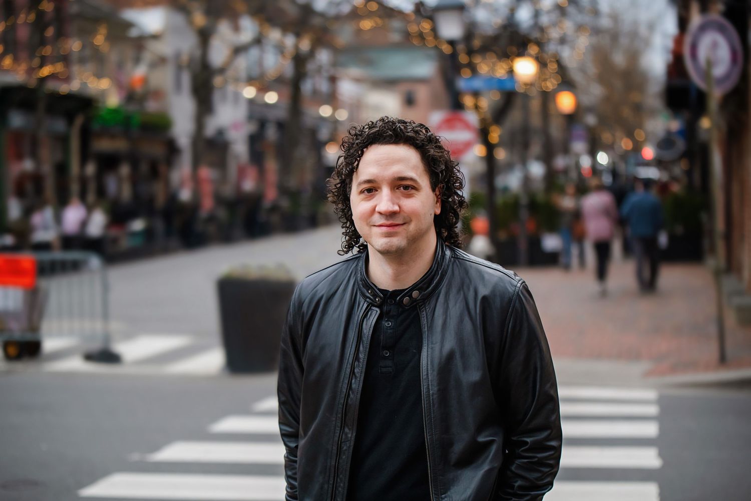 Person in black jacket standing on urban street with buildings and sidewalk in background.