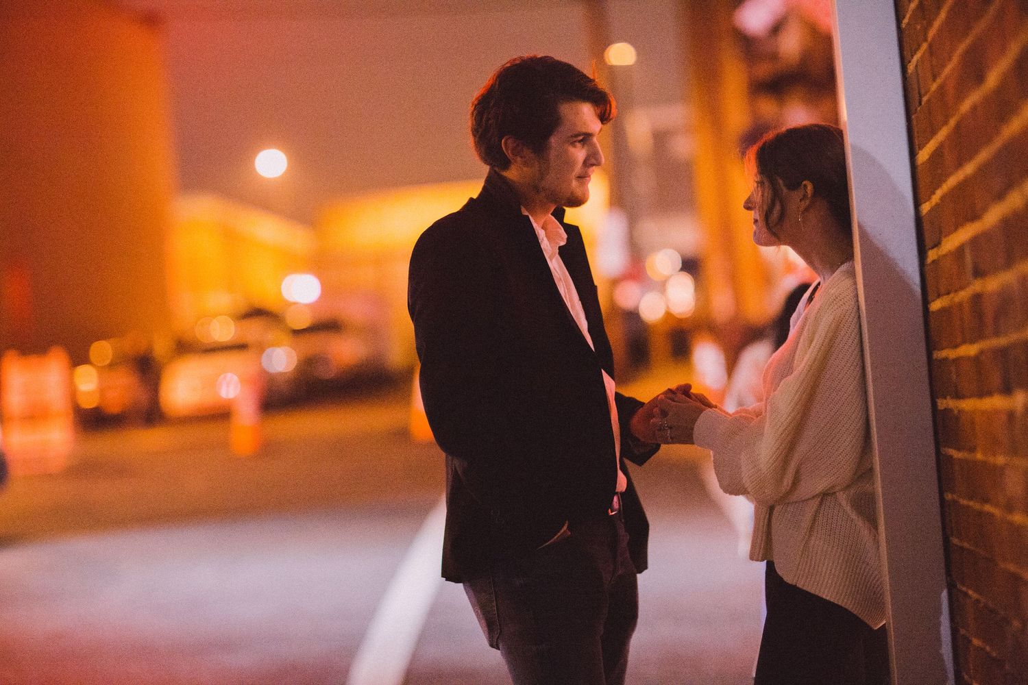 A couple shares an intimate moment on a city sidewalk at night under warm lighting.