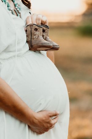 Mom to be in a white dress during golden hour holding her baby boots in Houston.