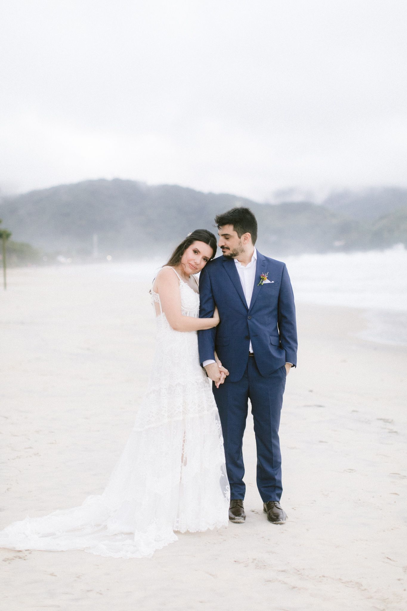 Couple in wedding attire walking along misty beach with mountains in background.