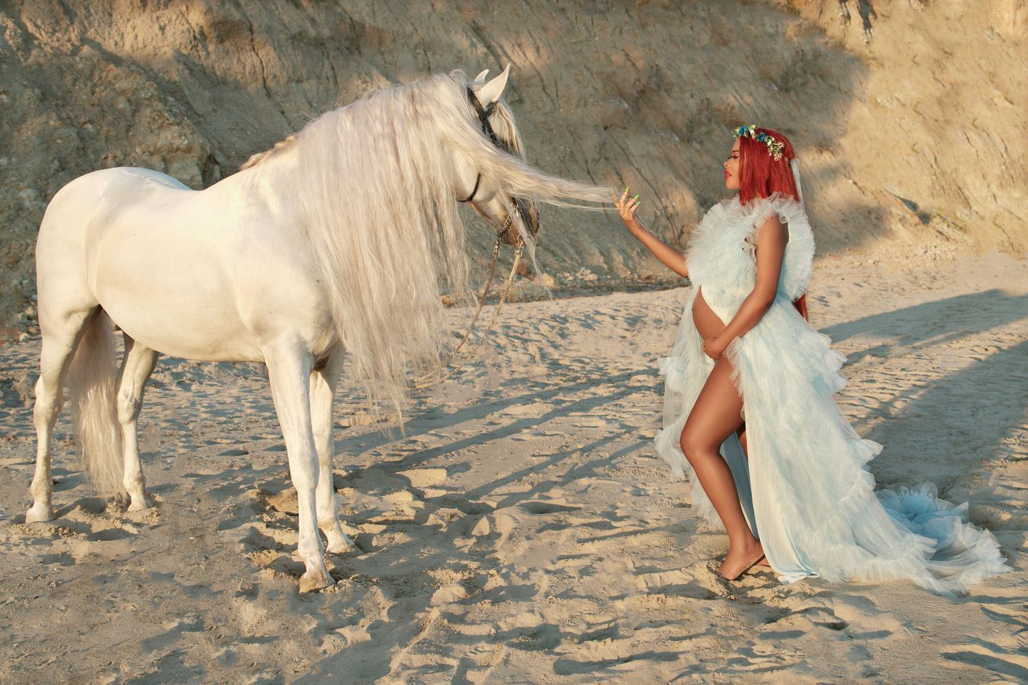 A dreamy scene with a white horse and figure in flowing dress on a sandy beach at sunset.