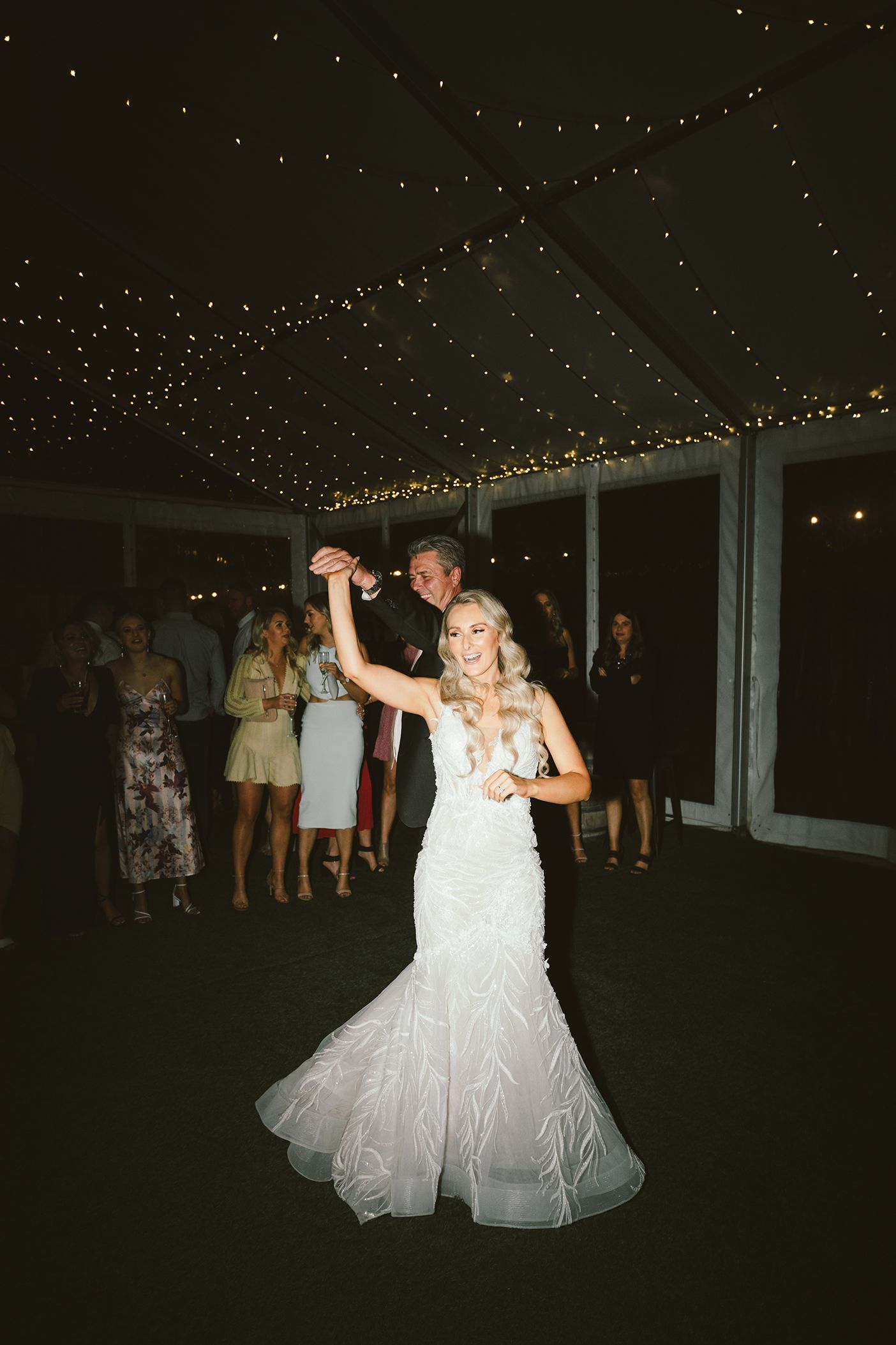 A bride in a white gown twirls on the dance floor under string lights at a wedding reception.