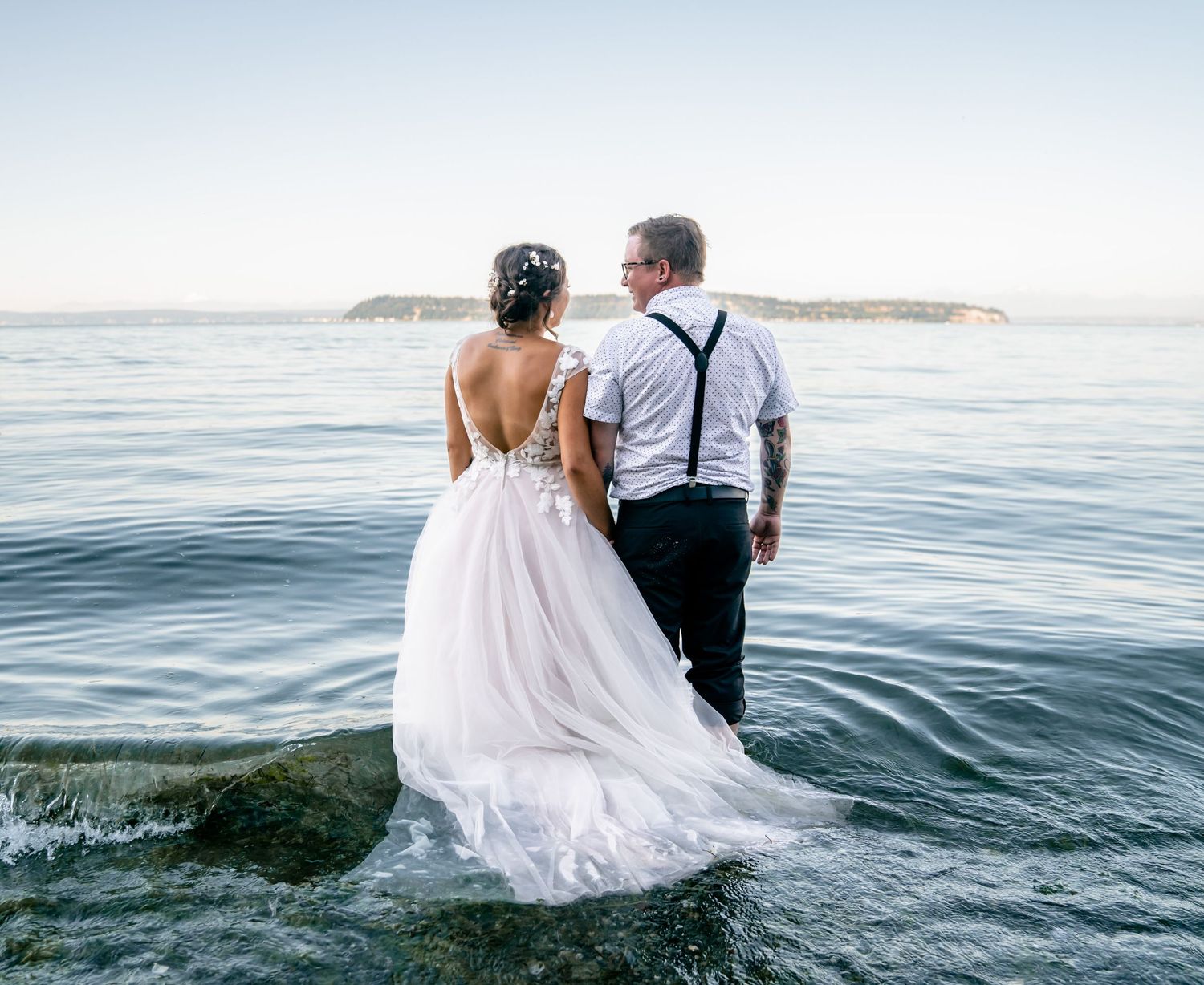Couple standing in ocean waves with white formal attire against serene seascape.