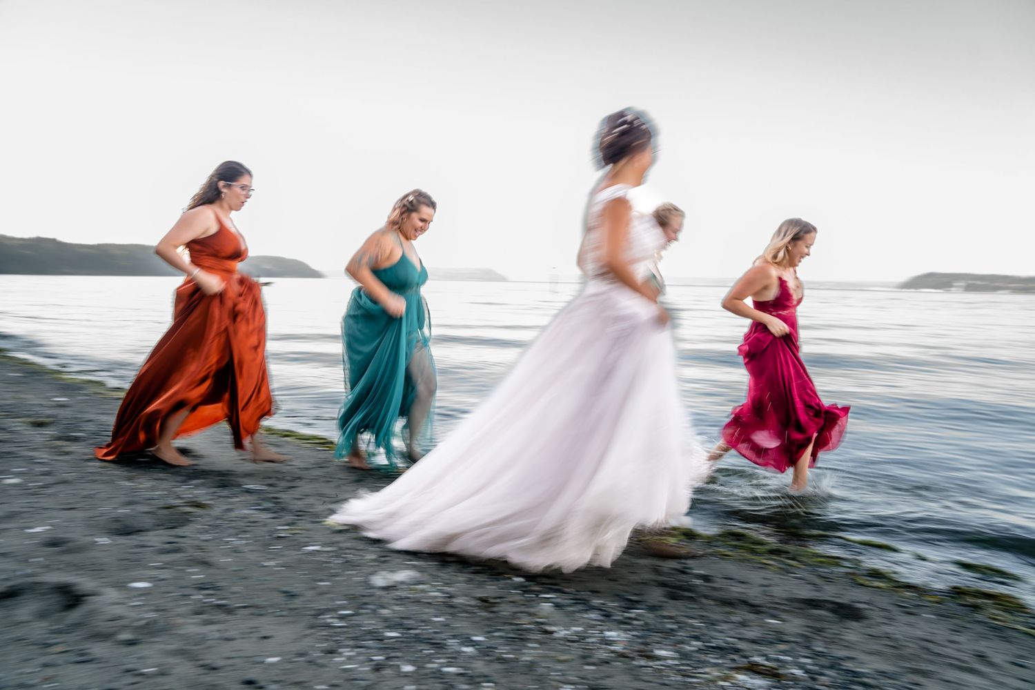 Group in colorful formal dresses walking through ocean waves at beach.