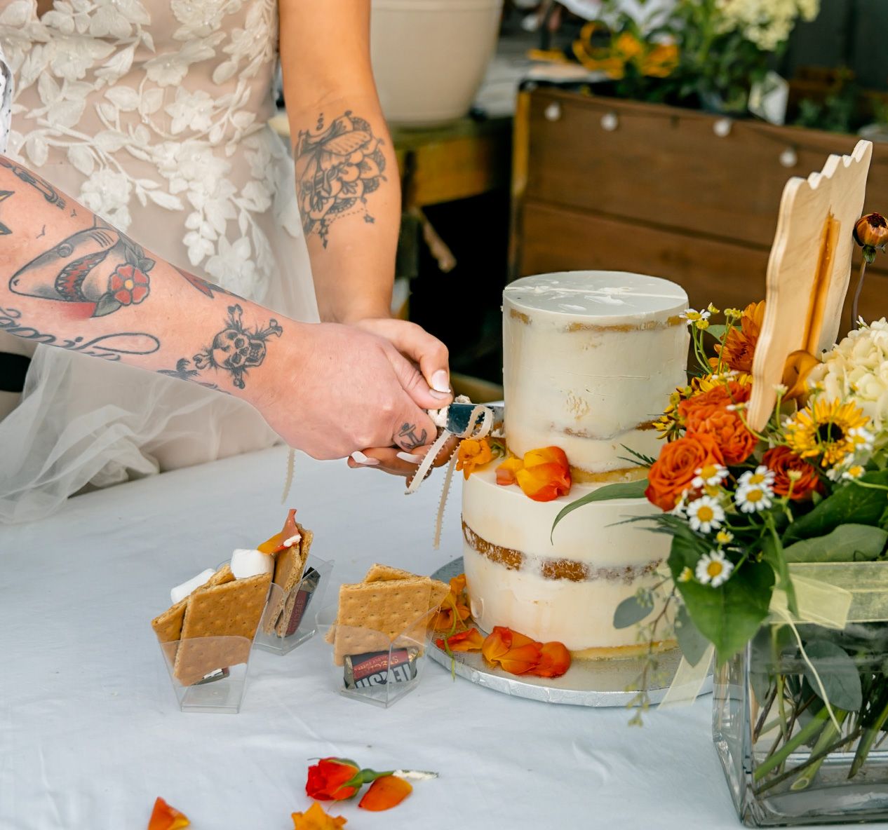 Tattooed hand cutting into a white wedding cake decorated with fresh flowers and orange accents.