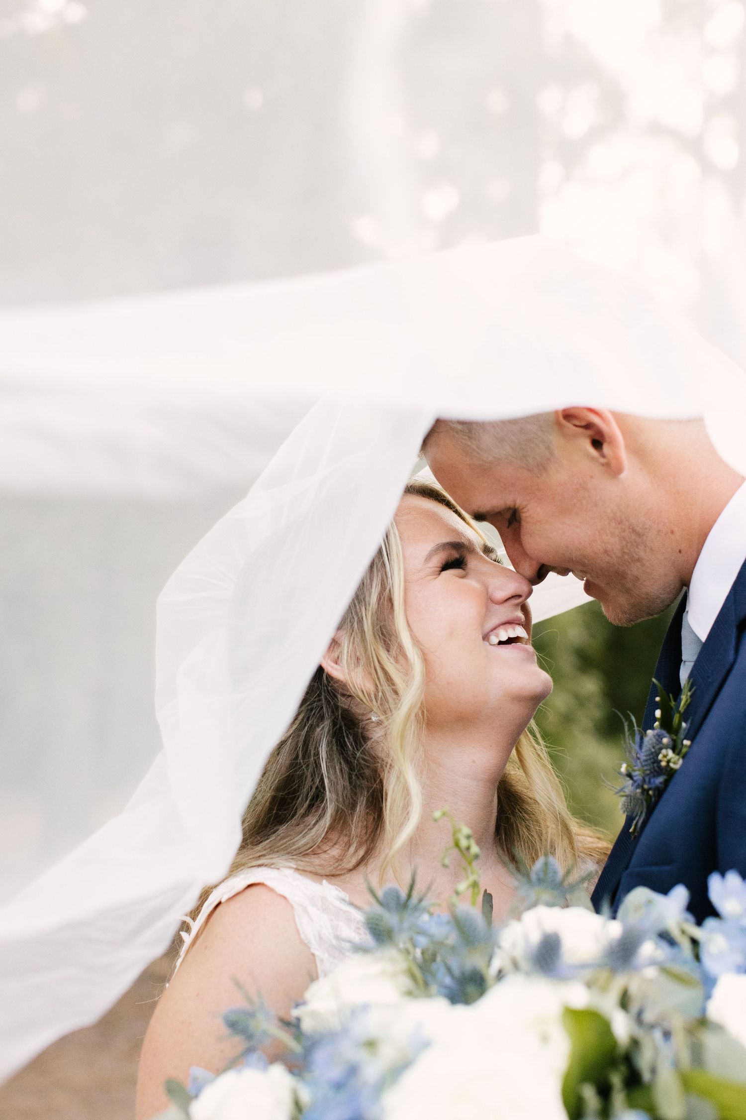 Romantic wedding moment captured under flowing veil with white and blue floral bouquet.