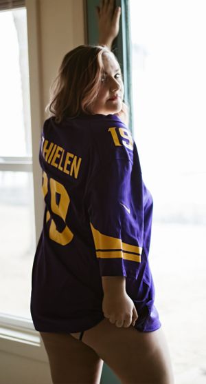 Woman wearing purple sports jersey poses by window in casual lifestyle portrait.